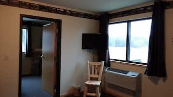 Quarto com cadeira de madeira, televisão, aparelho de ar condicionado e janela com cortinas.