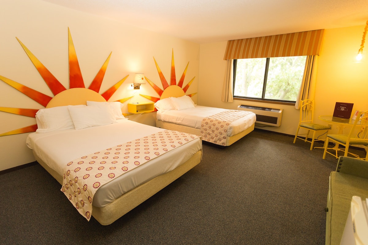 A hotel room with two beds, a sun-shaped wall decoration, a window with curtains, a small table, and a chair.