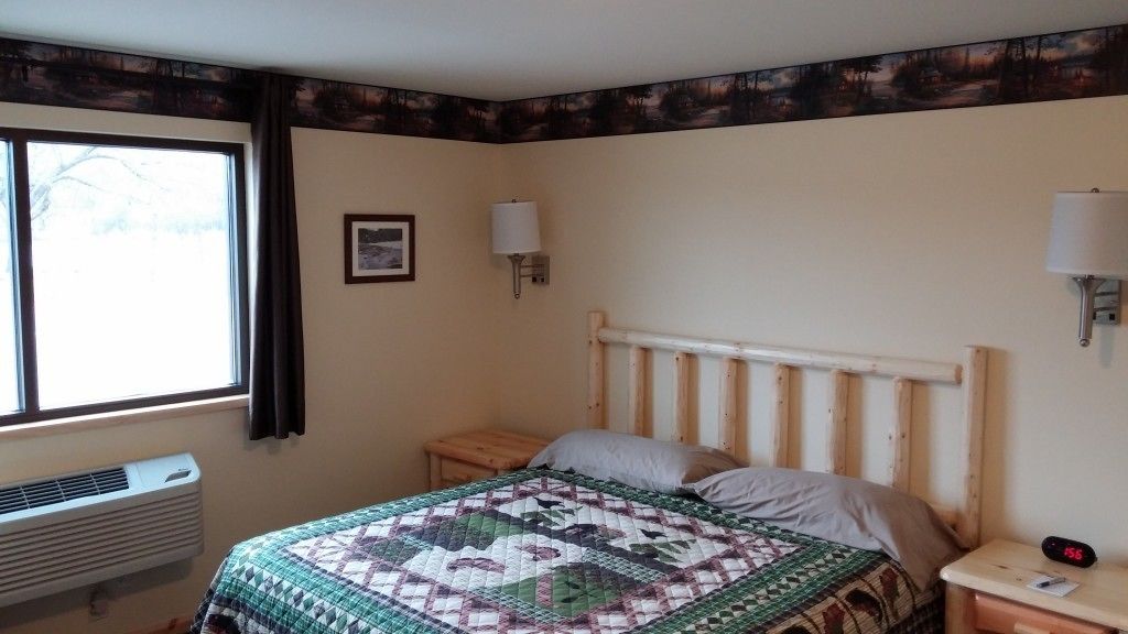 A bedroom with a wooden bed, a nightstand, a wall-mounted lamp, a window, and a radiator.