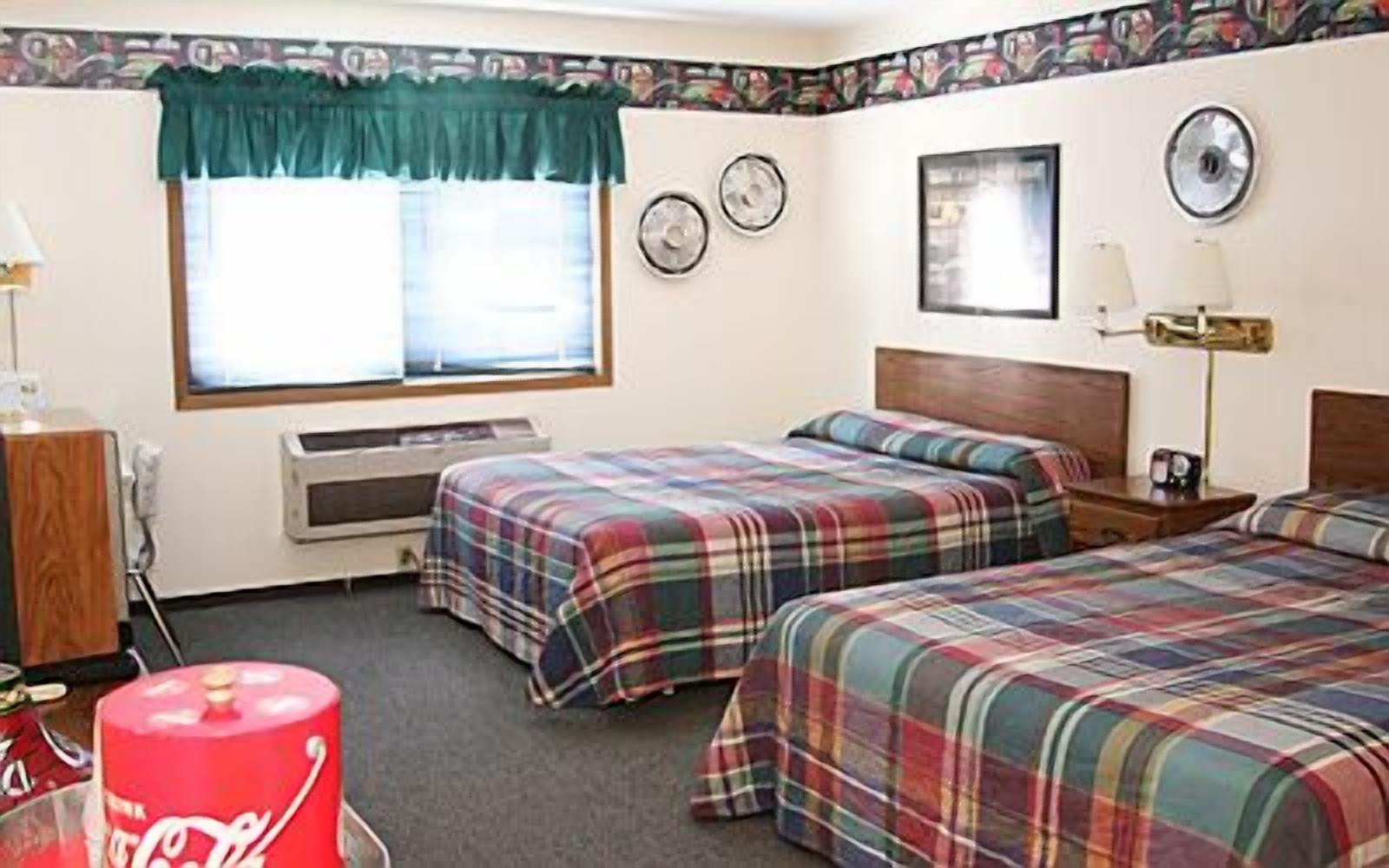 A motel room with two beds, a window with curtains, a microwave, a clock, and a wall-mounted lamp.