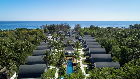 Un resort con varias edificaciones tipo bungaló dispuestas en fila, una piscina central y vista al océano.