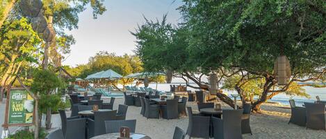 Een terras buiten met rieten tafels en stoelen, parasols en uitzicht op het strand.