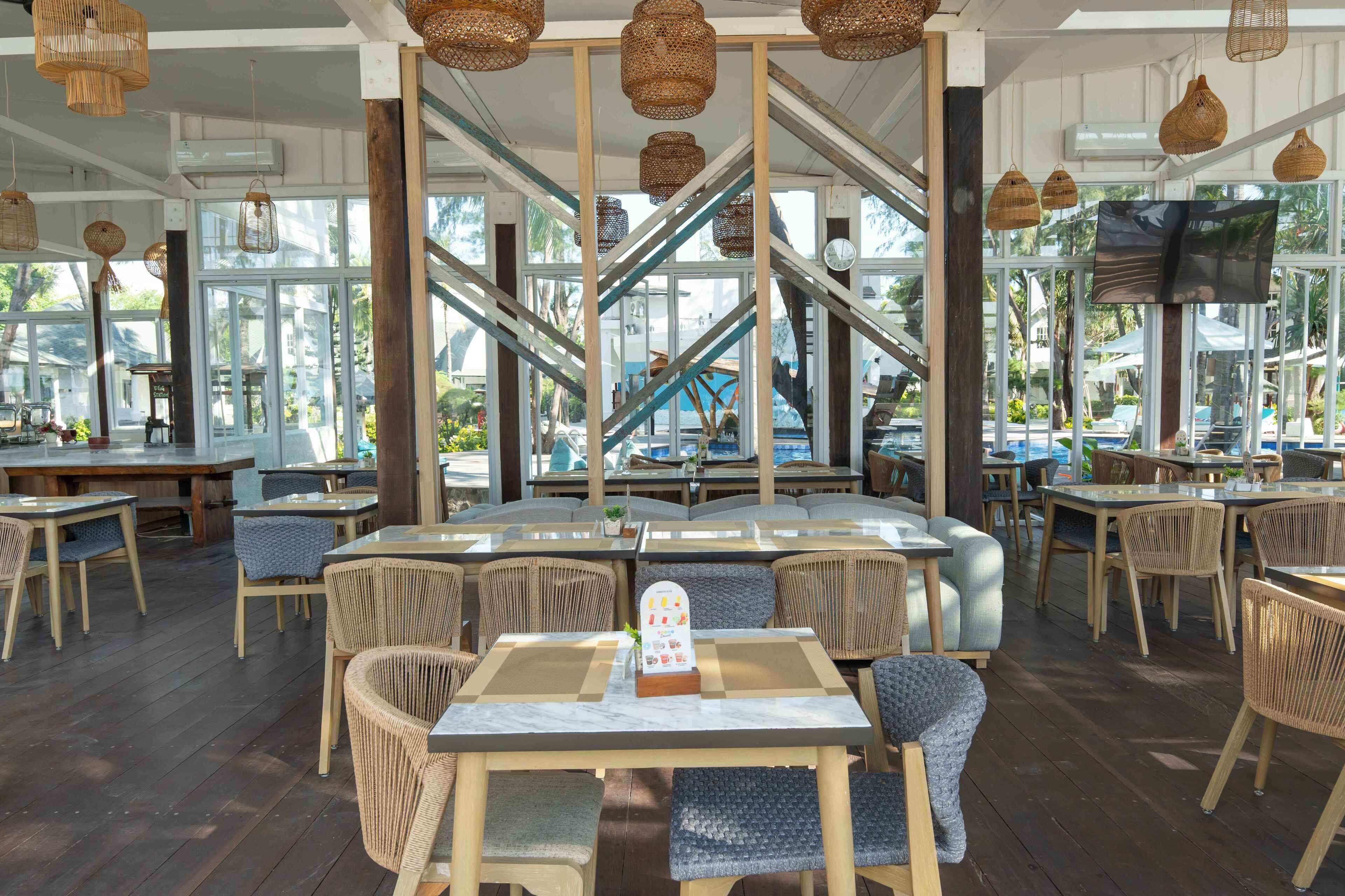 Uma área de refeições ao ar livre com mobiliário de vime, um bar central e grandes janelas com vista para uma área de piscina.