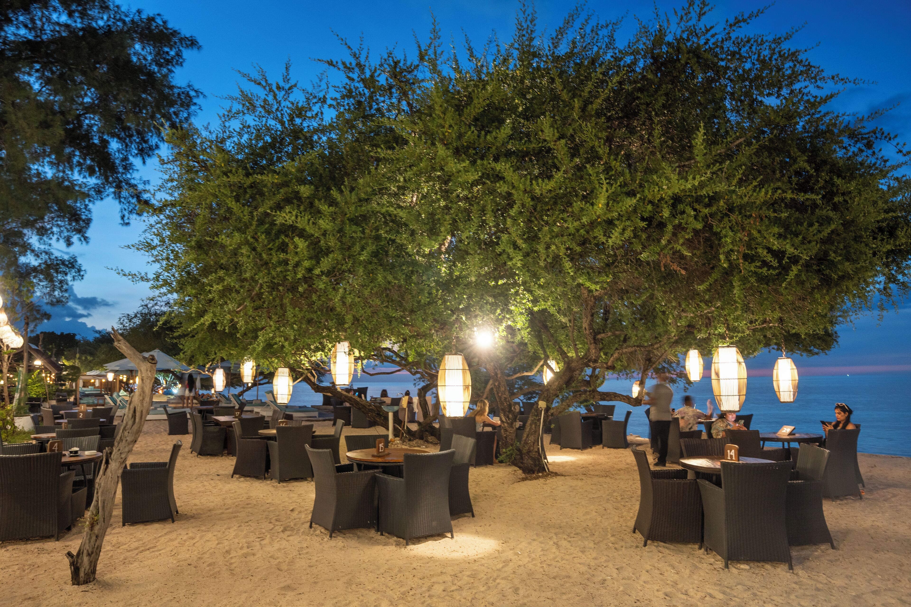An outdoor dining area with wicker furniture and hanging lights, overlooking a beach.