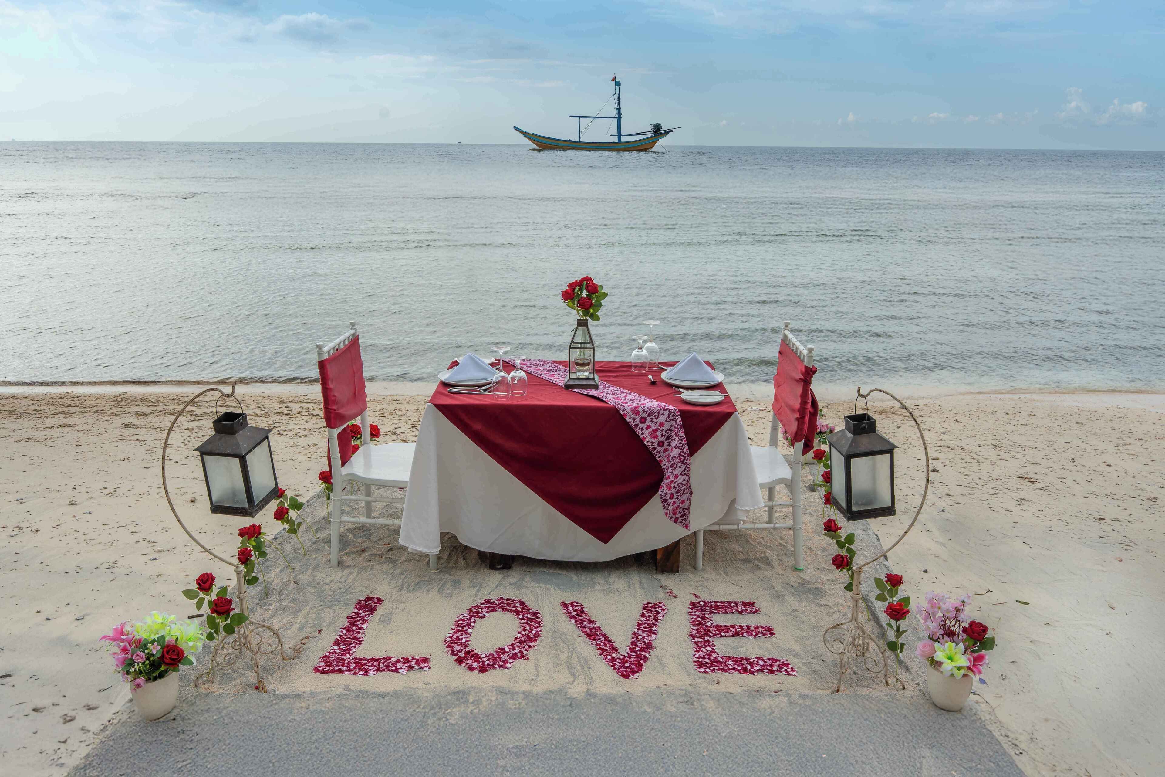 A romantic beach setting with a table set for two, adorned with red roses and a 'LOVE' sign on the sand.