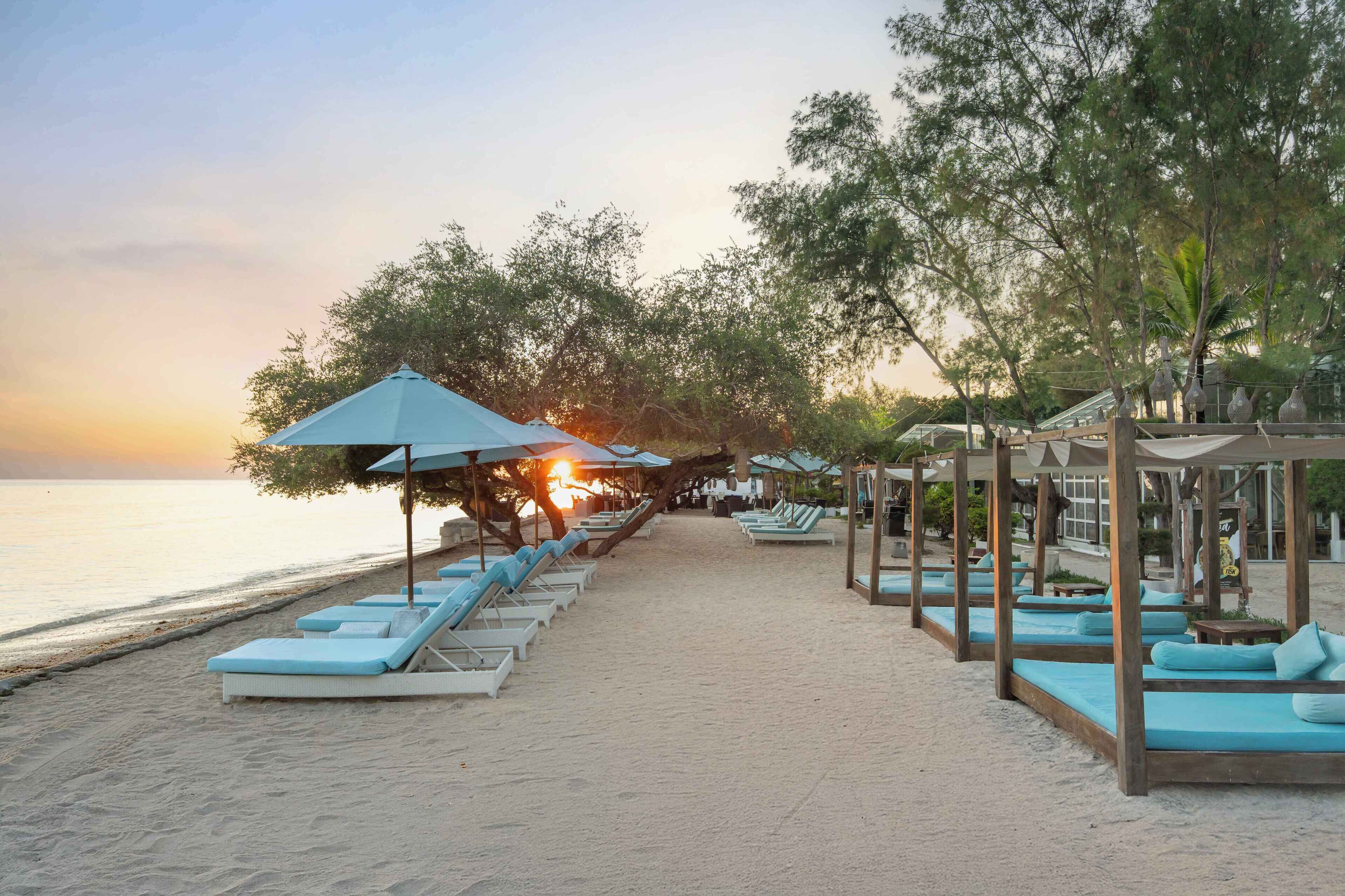 Un hôtel situé en bord de mer, doté de chaises longues et de parasols le long de la plage, d’un sentier menant à la plage et offrant une vue sur le coucher de soleil.