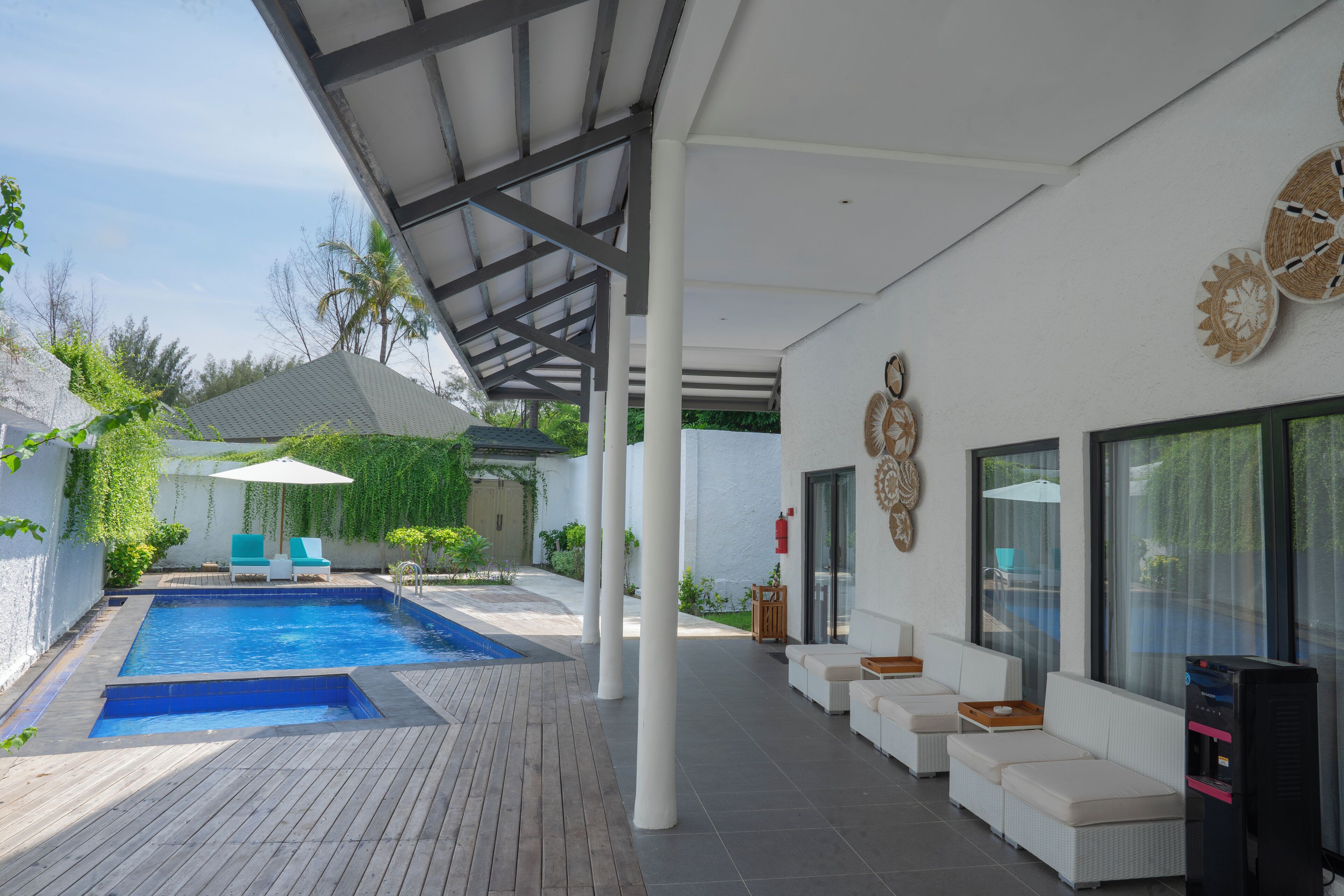 Un área de piscina moderna al aire libre con un espacio de estar techado, una terraza de madera y una pared blanca con detalles decorativos.