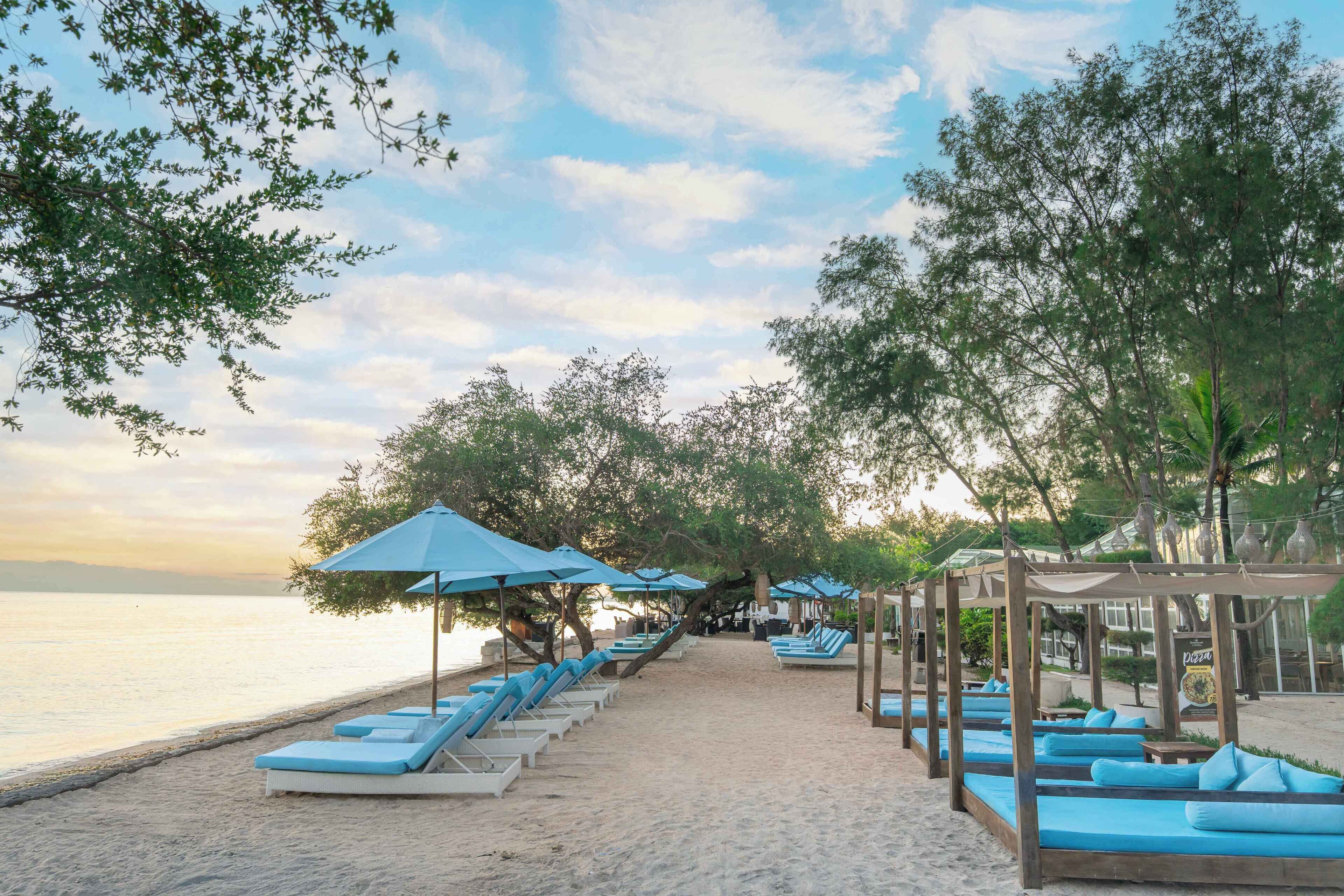 Un complexe hôtelier en bord de mer, avec des rangées de chaises longues et de parasols, une promenade bordée de sable et une vue sur la mer.