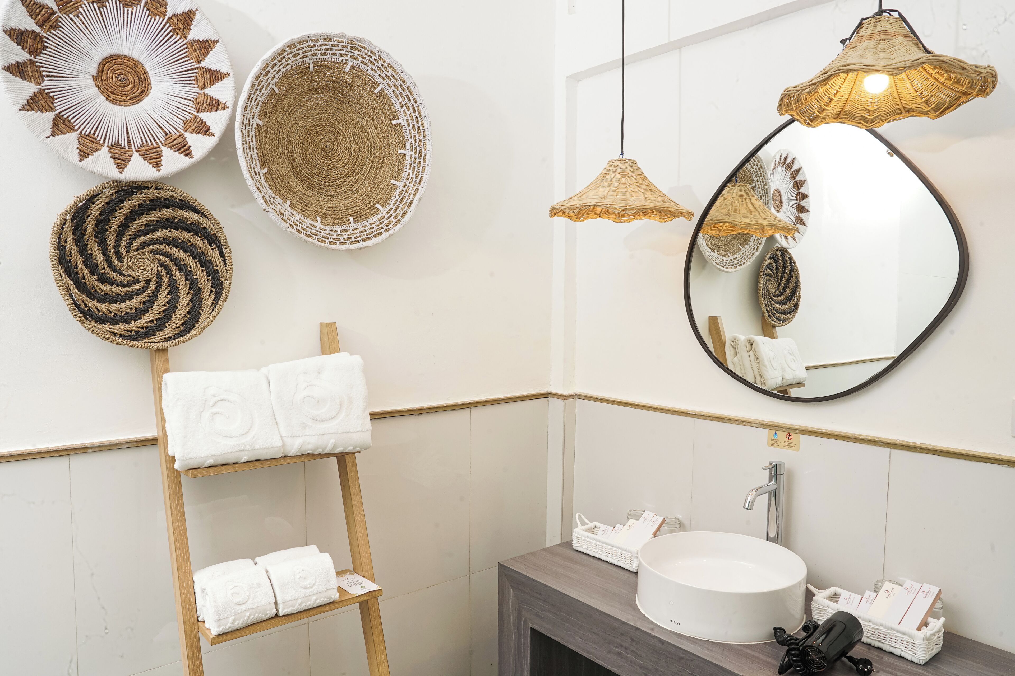 A bathroom with a round mirror, a wooden towel rack with towels, a round sink, and woven wall decorations.