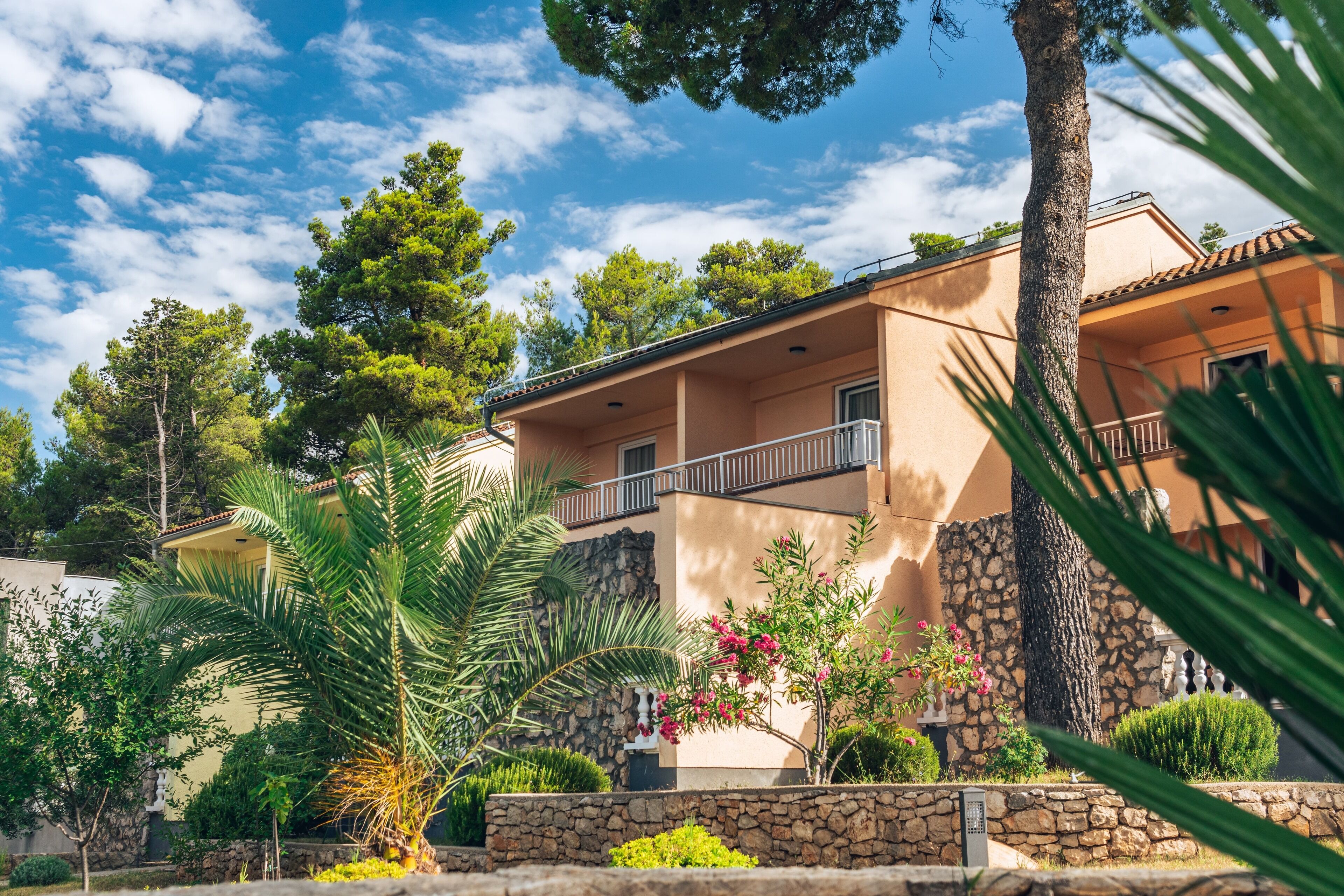 A two-story building with a stone facade, a balcony, and a well-maintained garden with palm trees and flowering plants.