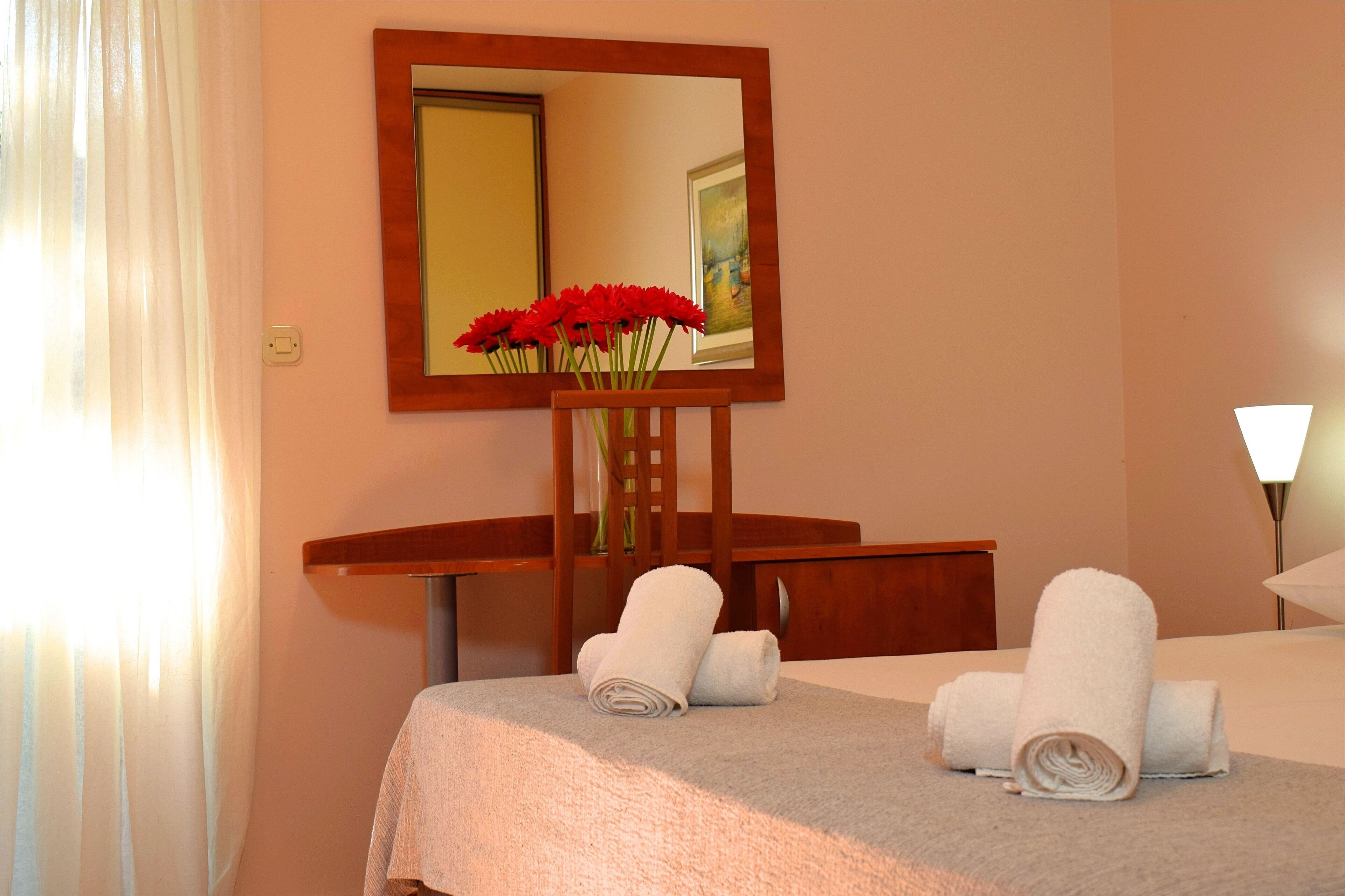 Quarto de hotel com uma cama, uma mesa de cabeceira de madeira com um vaso de flores vermelhas e um espelho na parede.