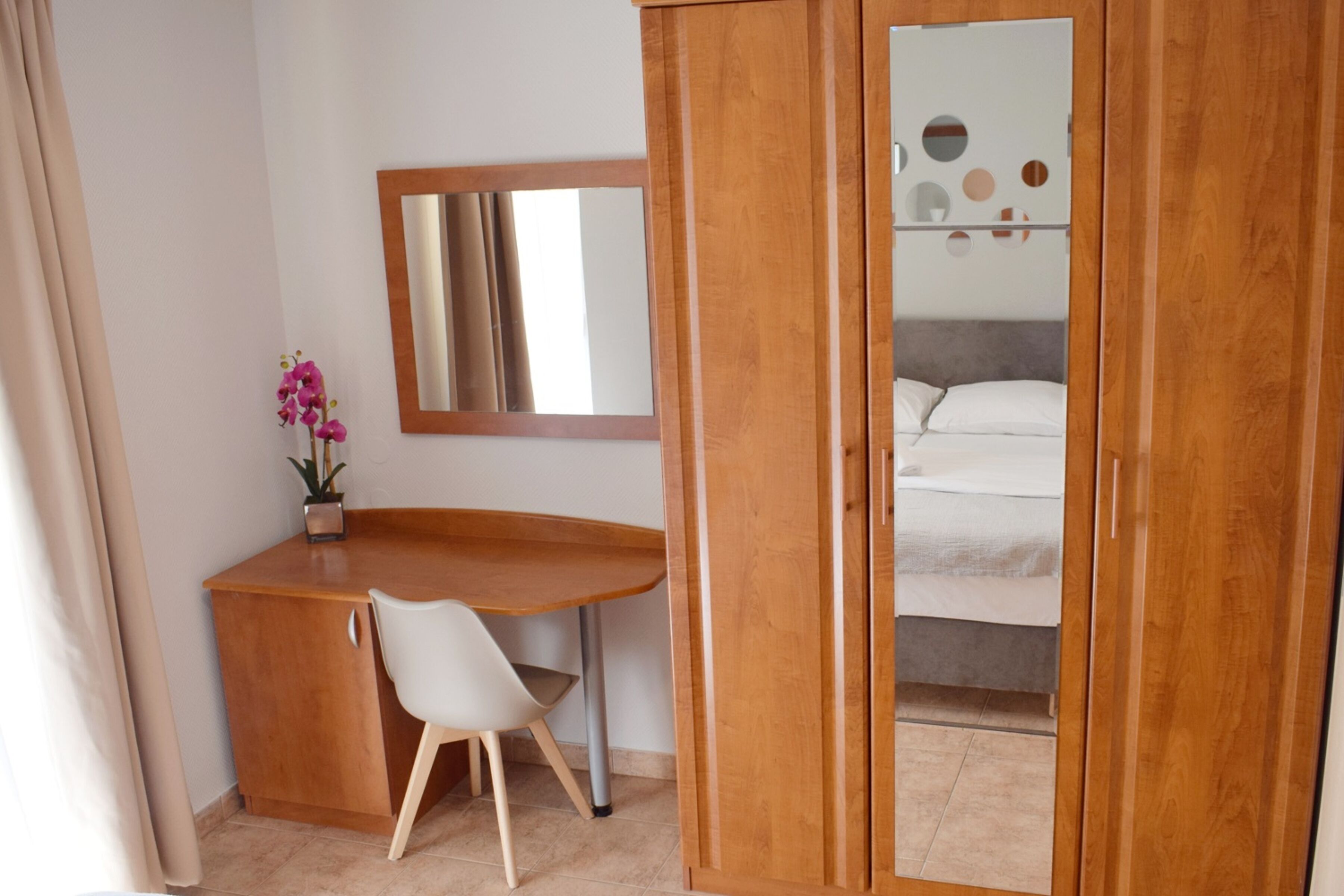 A room with a wooden desk, a chair, a bed with white bedding, a mirror, and a wall with circular decorations.
