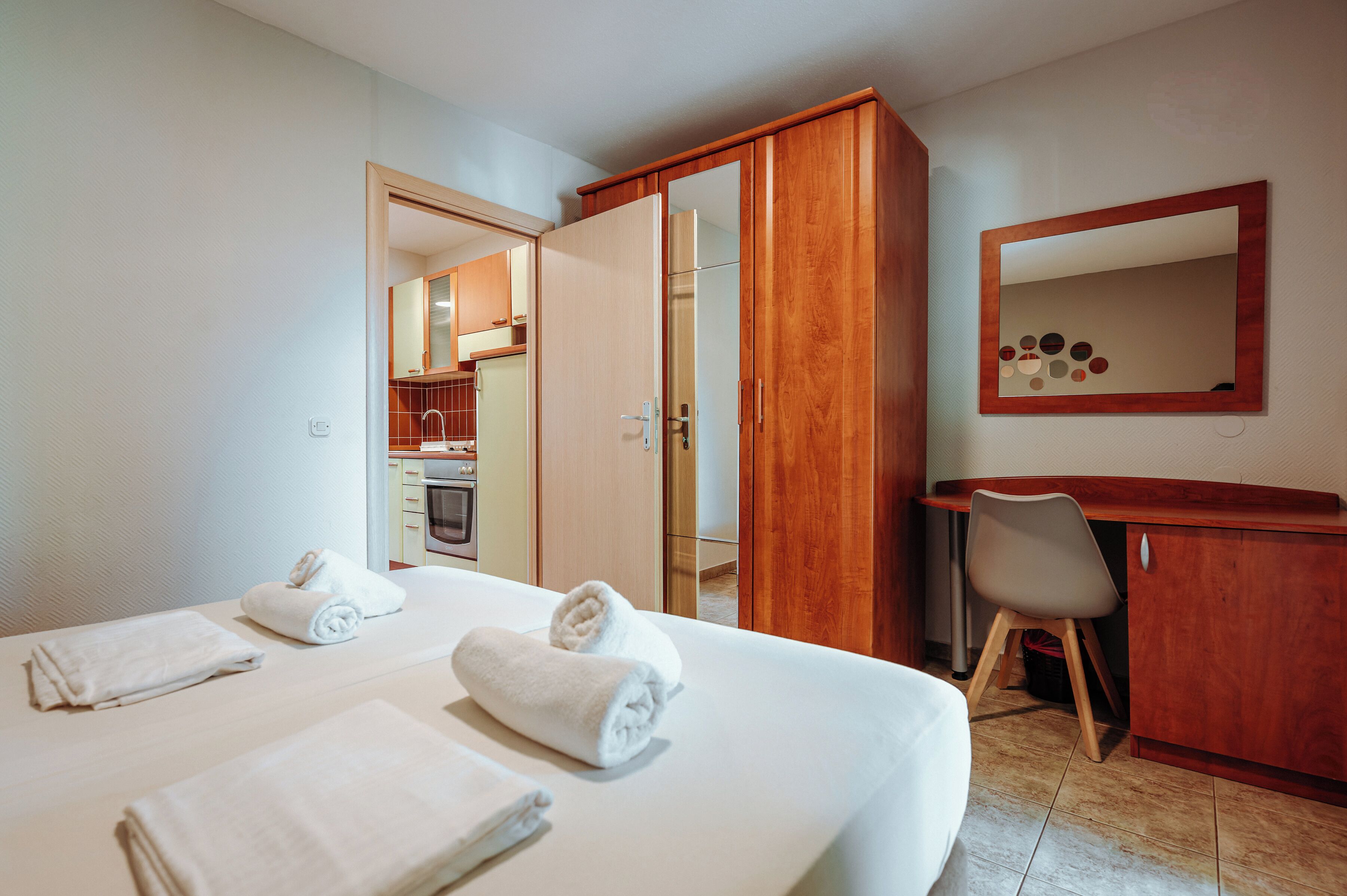 Camera d'albergo con un letto, due asciugamani, una scrivania, una sedia, uno specchio e una porta aperta che dà su un angolo cottura.