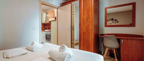 A hotel room with a bed, two towels, a desk, a chair, a mirror, and an open door leading to a kitchen area.