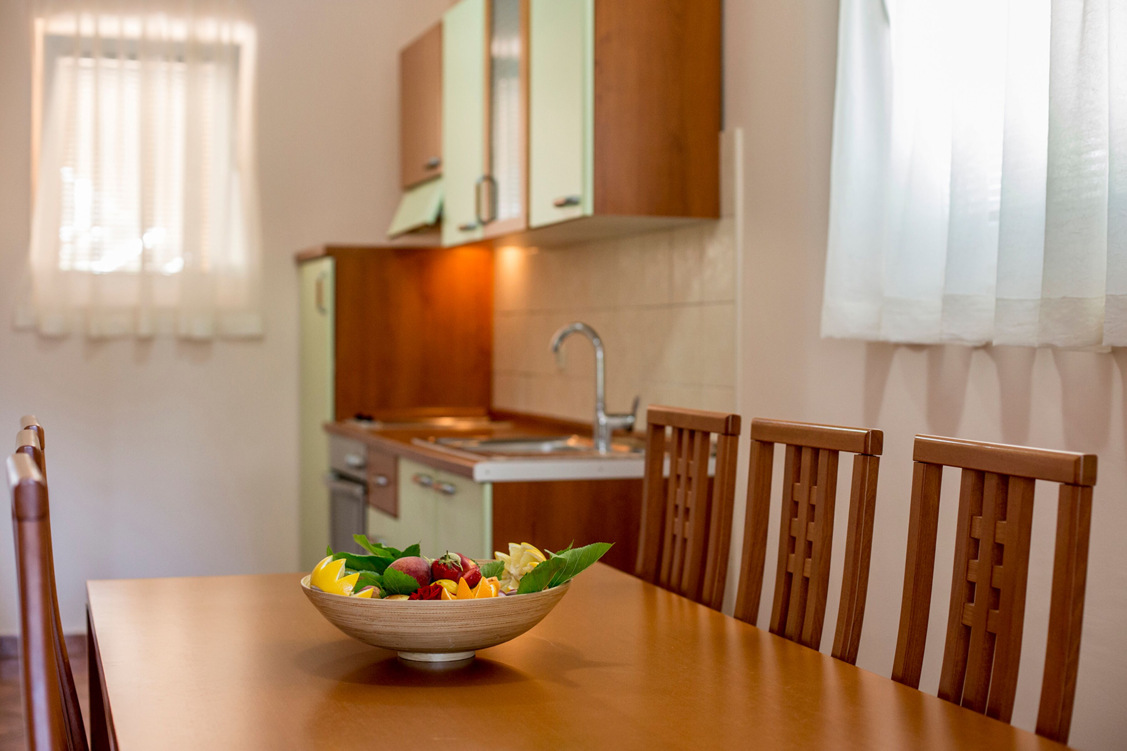 A dining table with a bowl of fruit, wooden chairs, a kitchen in the background, and a window with curtains.