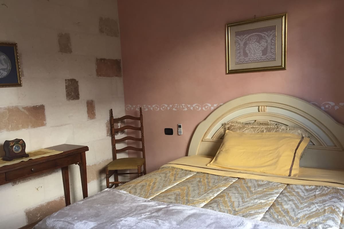 A bedroom with a bed, a wooden chair, a small table with a clock, and a framed picture on the wall.