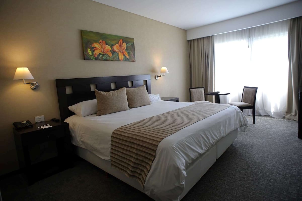 A hotel room with a large bed, two bedside tables, a chair, a painting, and a window with curtains.