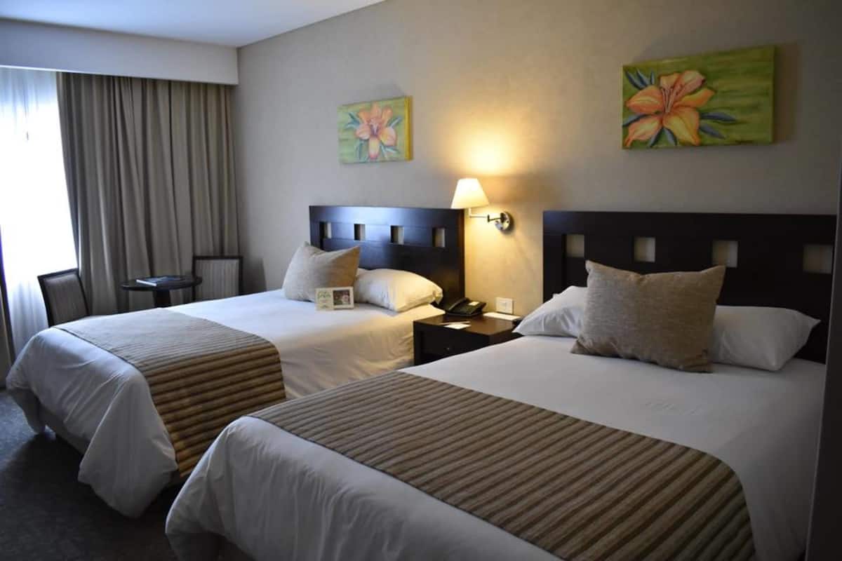 A hotel room with two beds, a bedside table with a lamp, and two paintings on the wall.