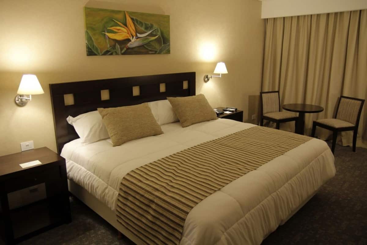 A hotel room with a large bed, two bedside lamps, a nightstand, a chair, a small table, and a painting on the wall.
