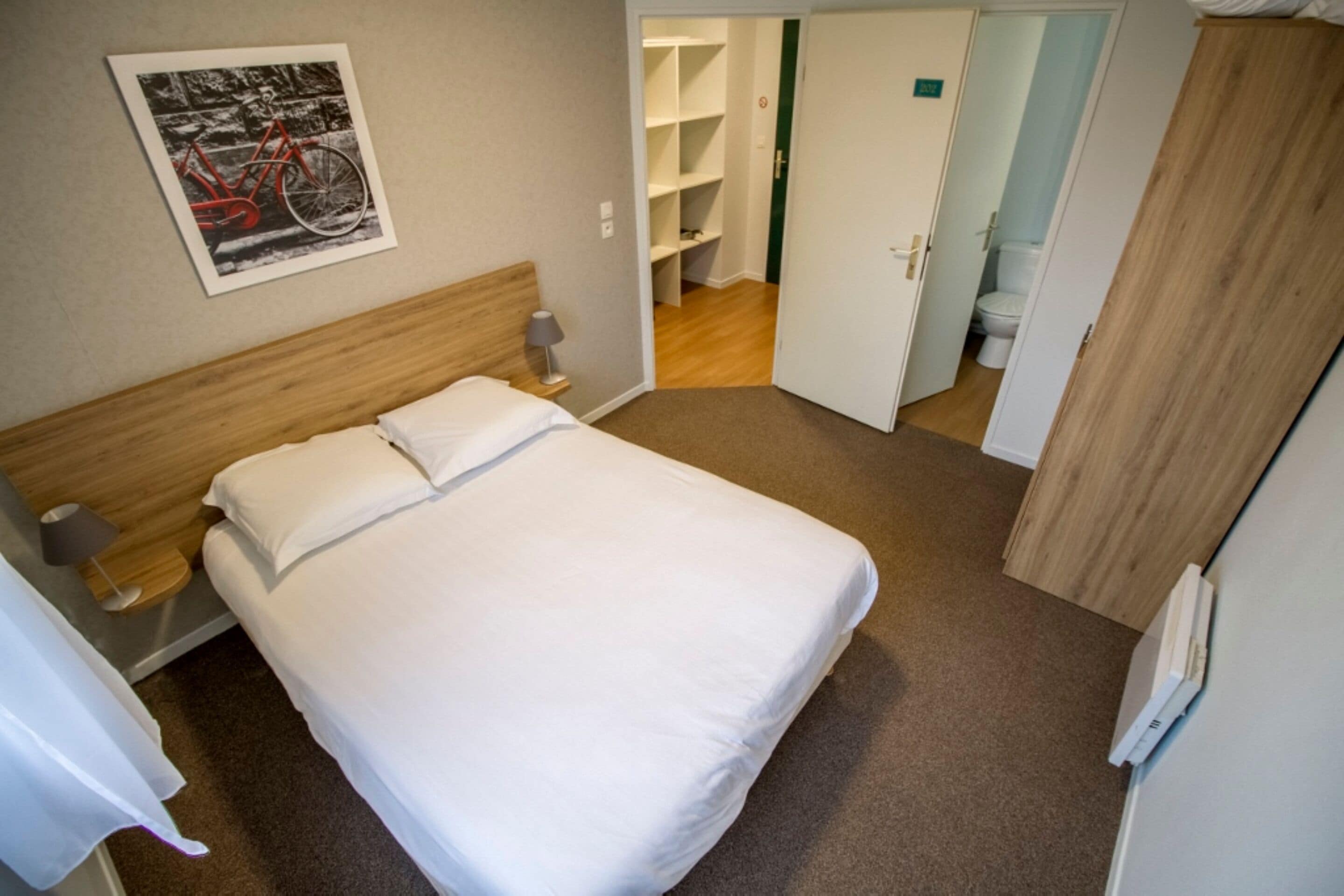 A hotel room with a single bed, a nightstand, a lamp, a framed picture on the wall, and a wooden wardrobe.