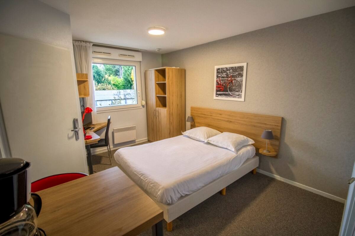 Ein Hotelzimmer mit Bett, Schreibtisch, Stuhl, Kleiderschrank und einem Fenster mit Blick ins Grüne.