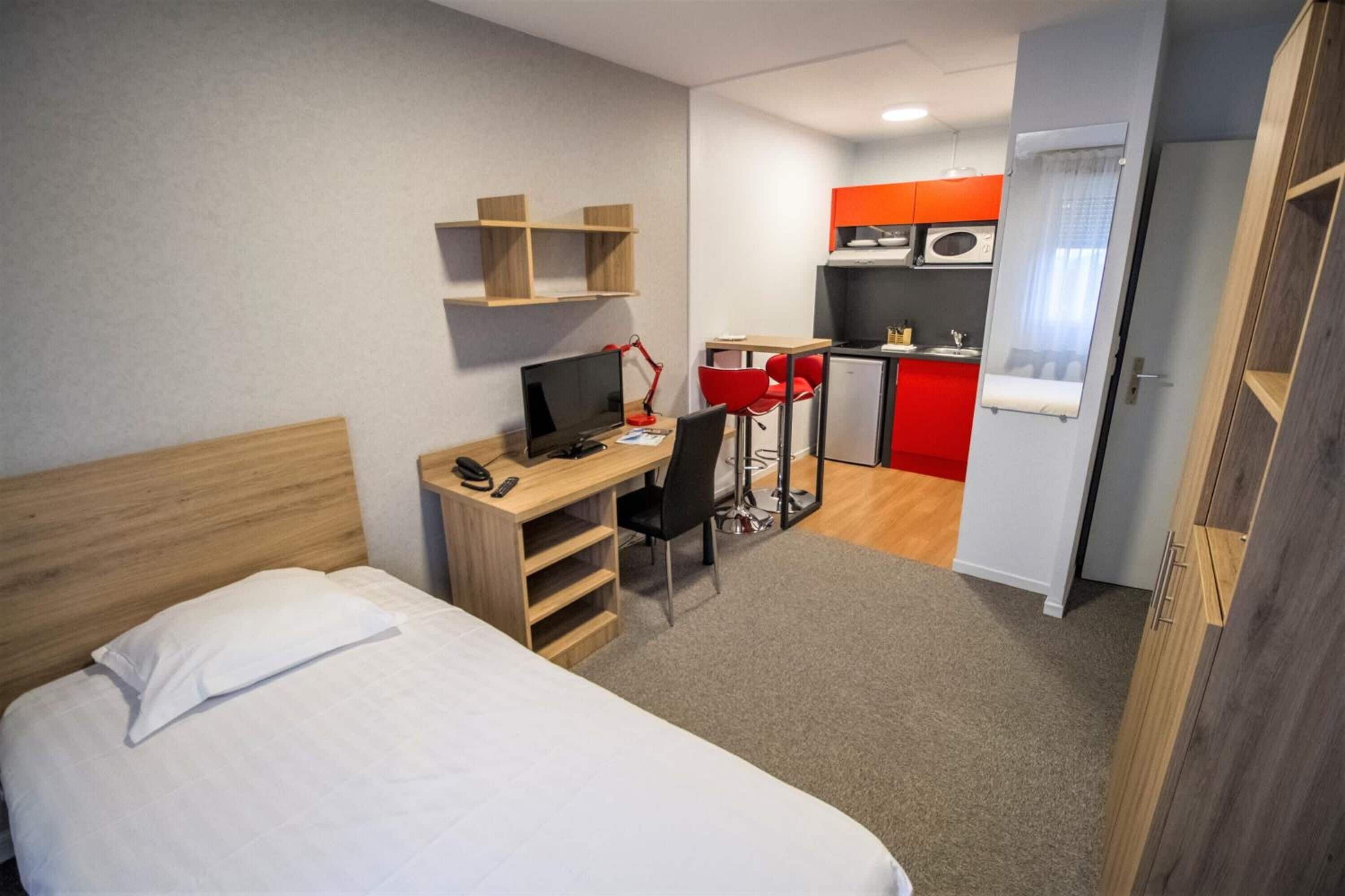 Une chambre d’hôtel comprenant un lit, un bureau avec un ordinateur, une chaise, une petite kitchenette et une porte.