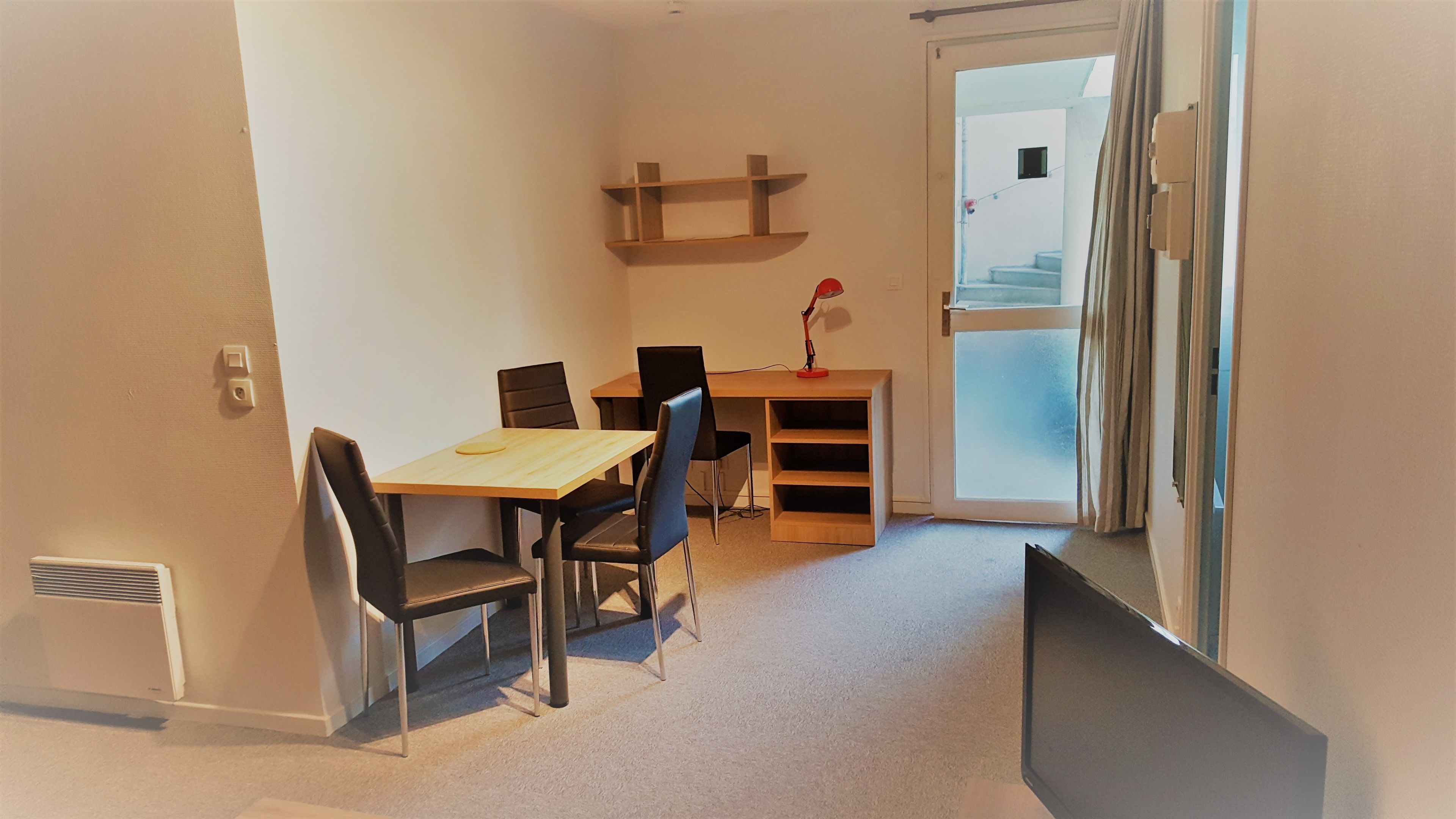 A small, well-lit room with a wooden desk, two black chairs, a small shelf, and a door leading to a balcony.