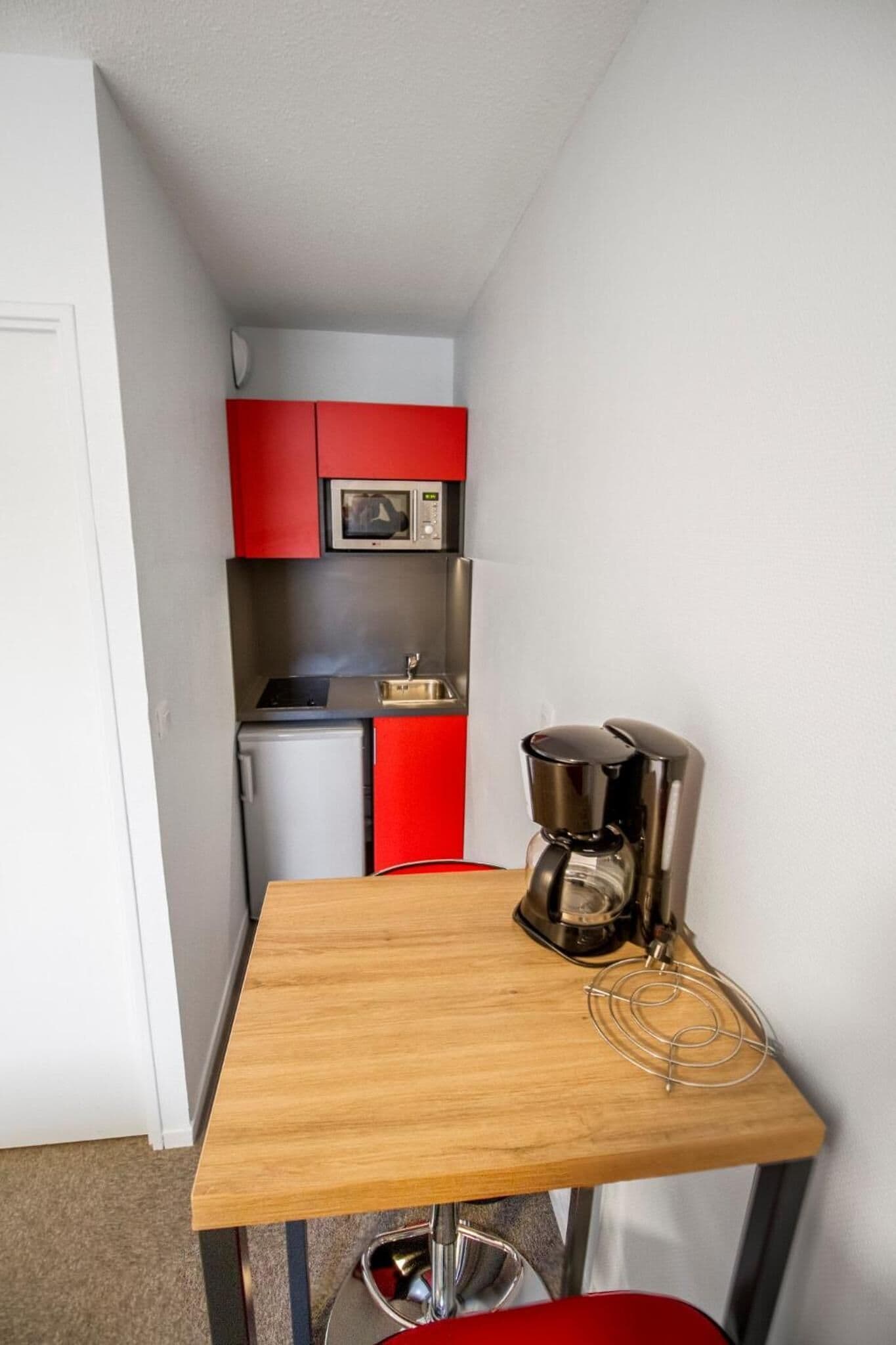 A small kitchenette with a microwave, sink, and red cabinets. There is a wooden table with a coffee maker and a red chair.