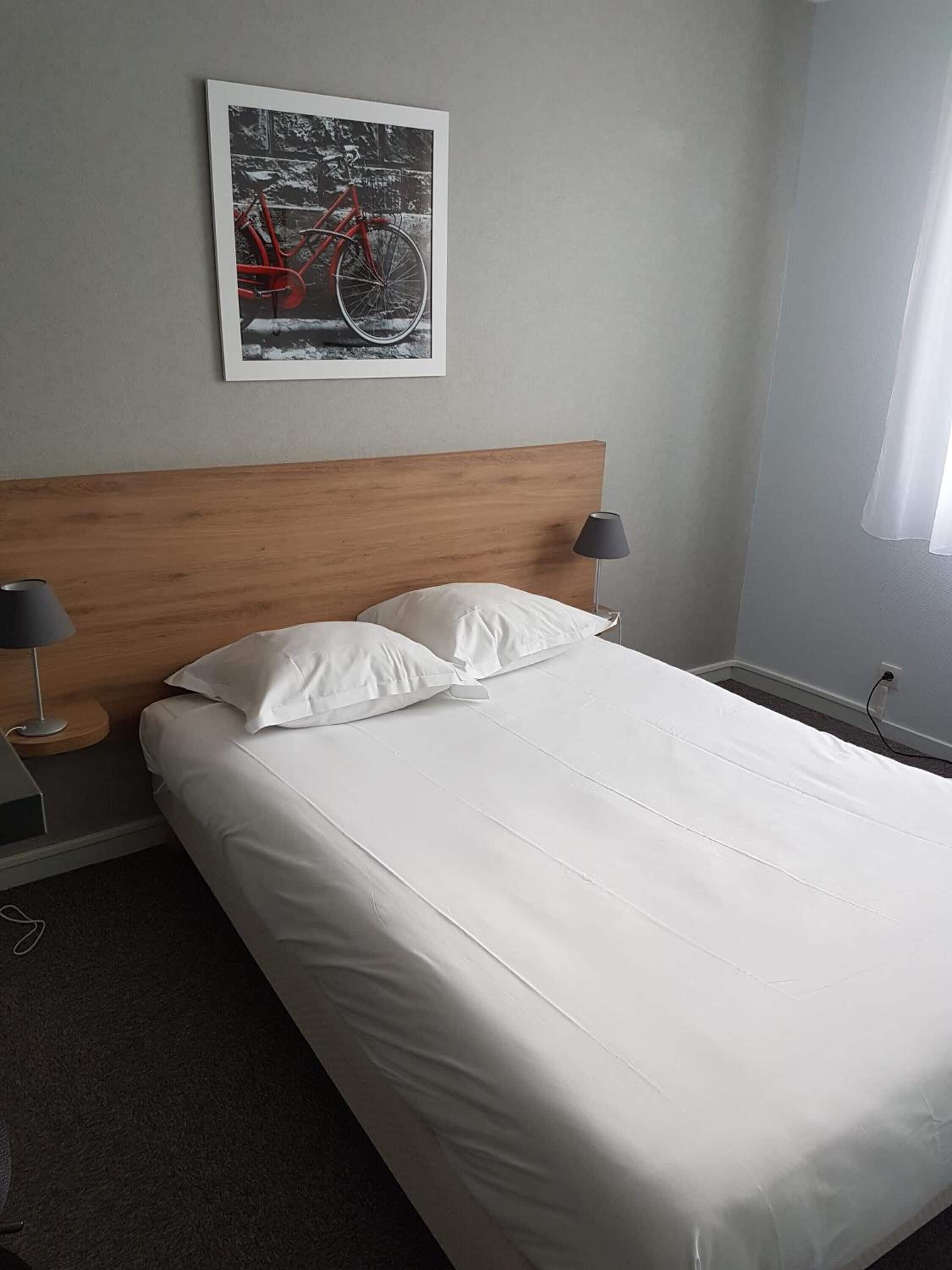 A neatly made bed with white linens in a room with a wooden headboard. There is a framed picture of a bicycle on the wall.