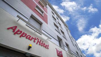 La façade d’un hôtel affiche en grandes lettres l’inscription « Appart Hotel ». Elle est décorée de couleurs rouge et blanche, et un ciel bleu clair, parsemé de nuages, se détache en arrière-plan.