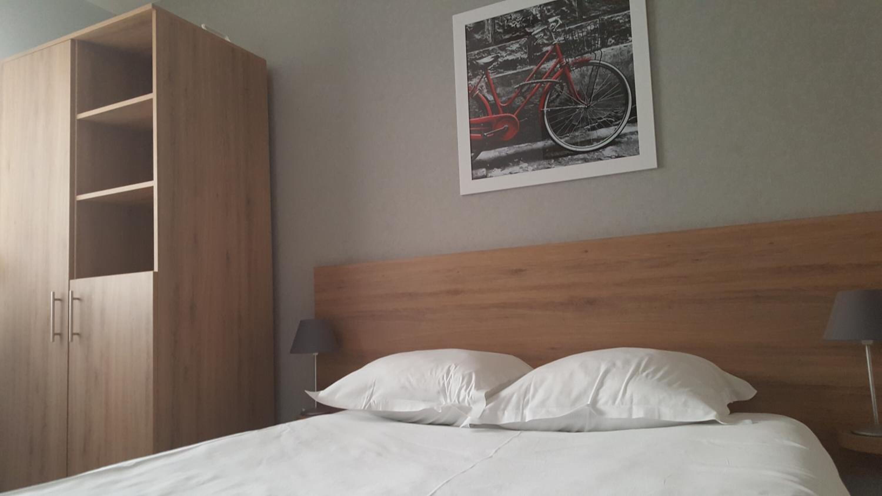 A hotel room with a wooden headboard, a bed with white linens, a large wardrobe, and a framed picture of a bicycle on the wall.