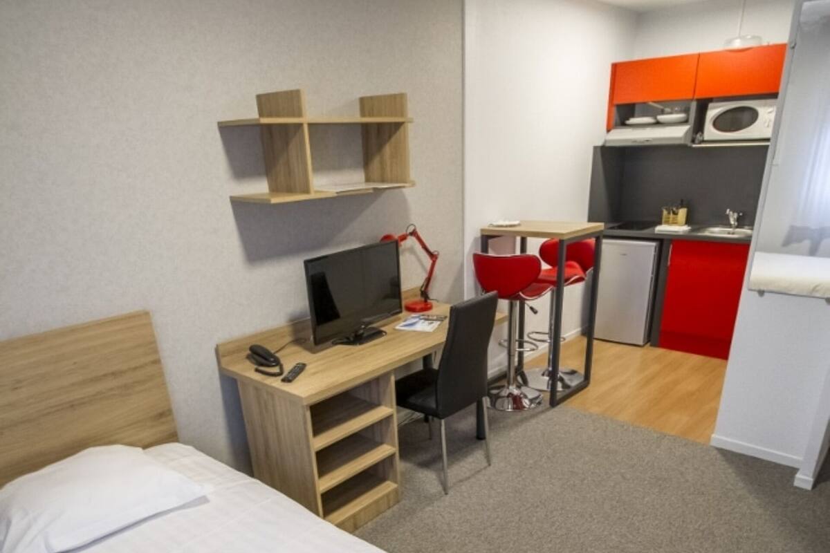 Ein Hotelzimmer mit einem Bett, einem Schreibtisch mit Computer, einem Sessel und einer kleinen Küche im Hintergrund.