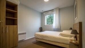 Een slaapkamer met een bed, een kledingkast, een raam met gordijnen, een nachtkastje met een lamp en een radiator.