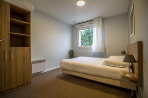 Une chambre à coucher comprenant un lit, une armoire, une fenêtre avec des rideaux, une table de chevet avec une lampe et un radiateur.