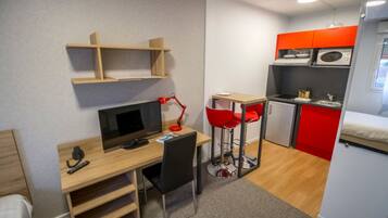Una habitación de hotel compacta con escritorio de madera, televisor, silla roja, kitchenette y cama.