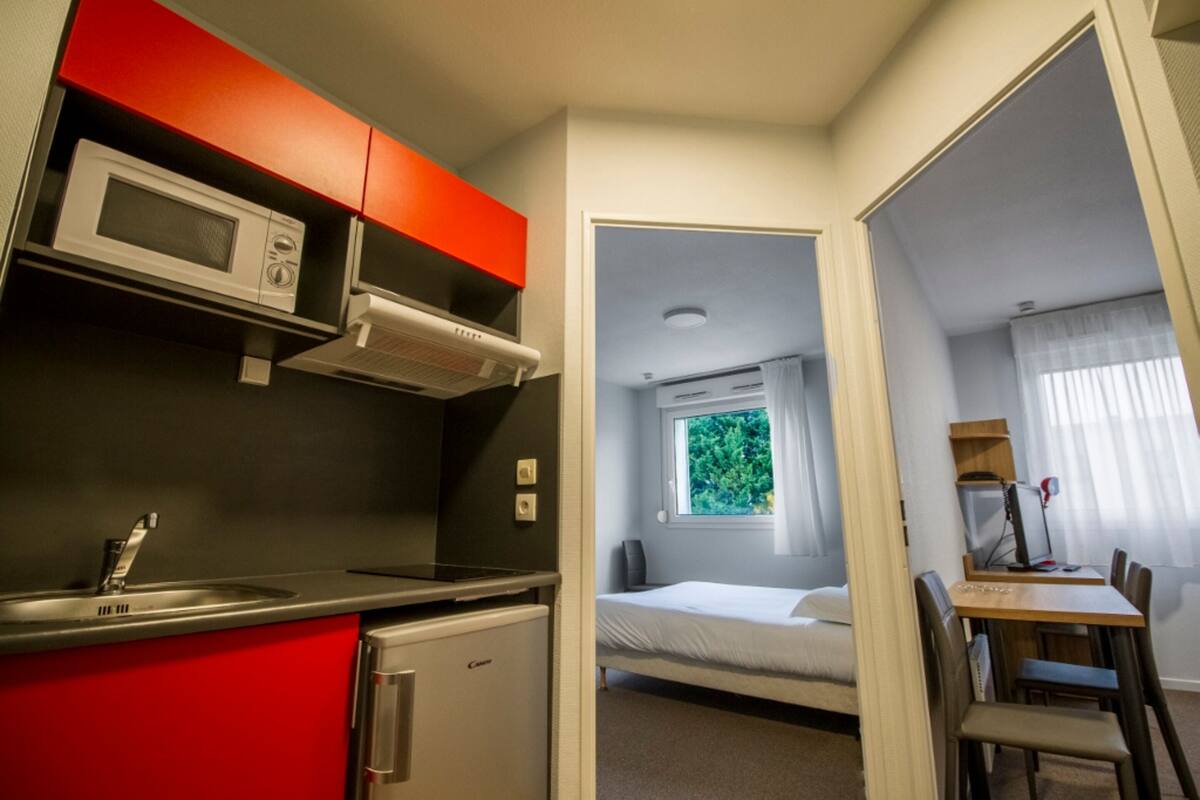 Ein gemütlicher Raum mit Küchenzeile, einem Bett, einem Schreibtisch und einem Fenster mit Blick auf Grünflächen.