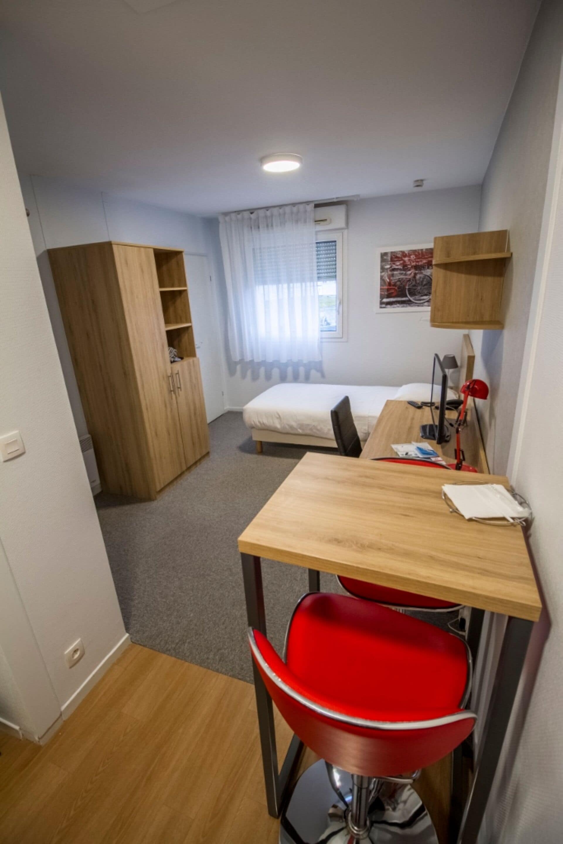 A compact room with a wooden desk, a red chair, a bed, a wardrobe, and a small kitchenette area.