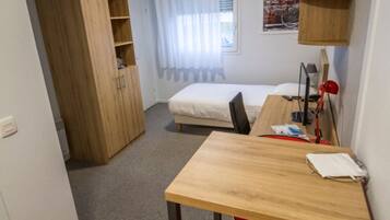 A compact room with a wooden desk, a red chair, a bed, a wardrobe, and a small kitchenette area.