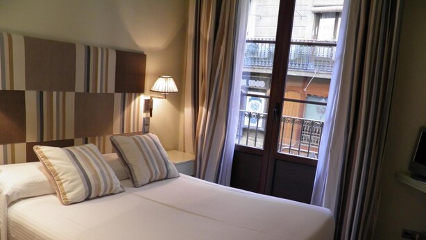A hotel room with a bed, two pillows, a nightstand with a lamp, a window with curtains, and a view of a balcony. - Hotel Annex (Barcelona)