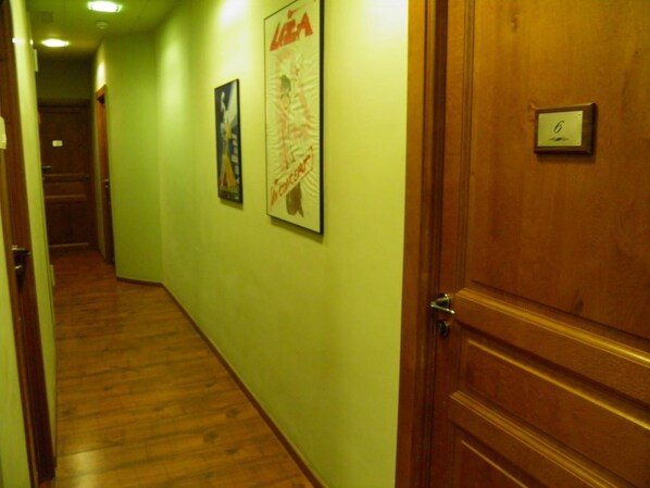 A hallway with wooden flooring, green walls, and two framed posters. A wooden door with a number plate is visible.