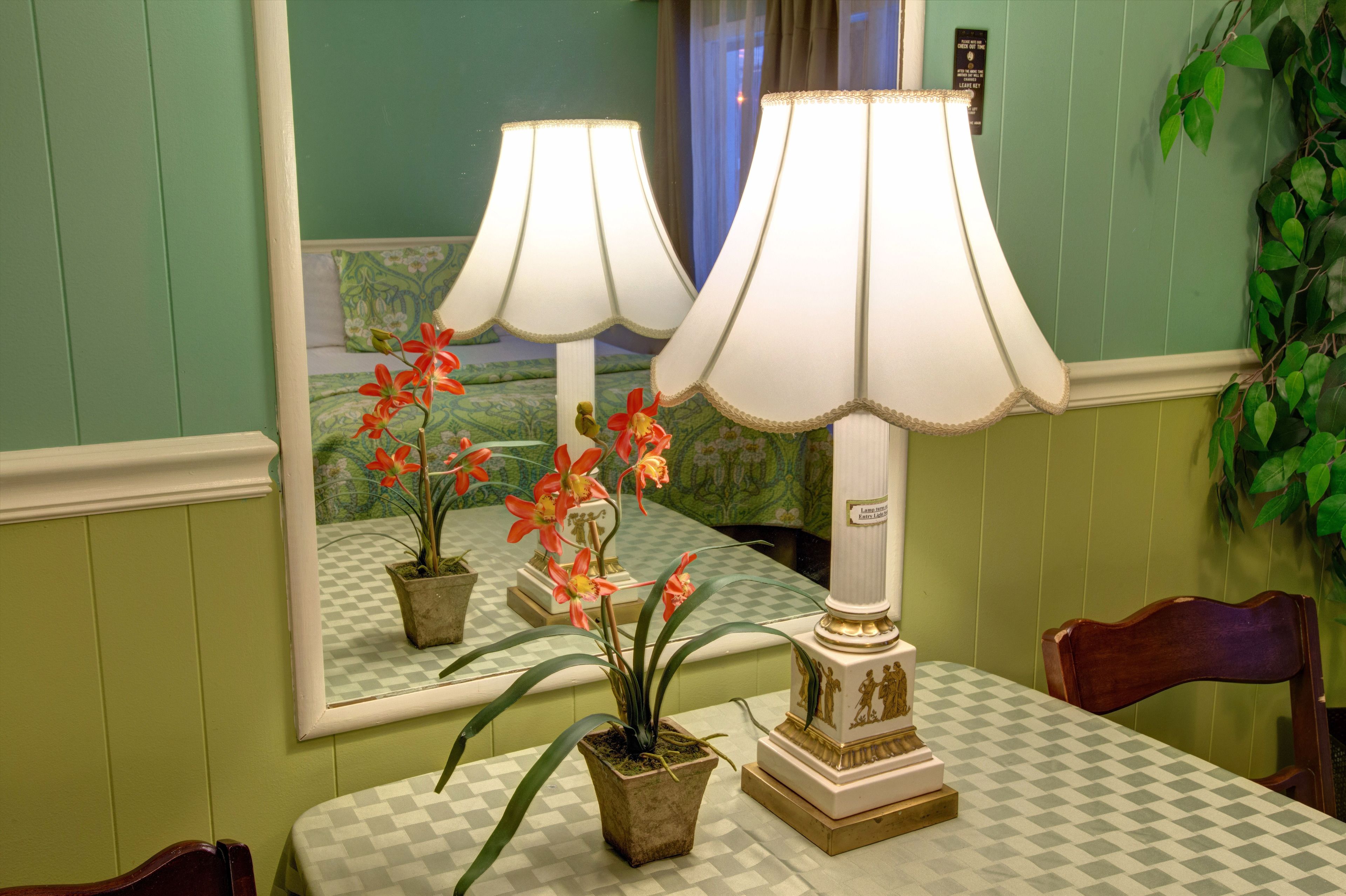 Una habitación con mantel a cuadros, una lámpara y un espejo que refleja una cama y una ventana con cortinas.