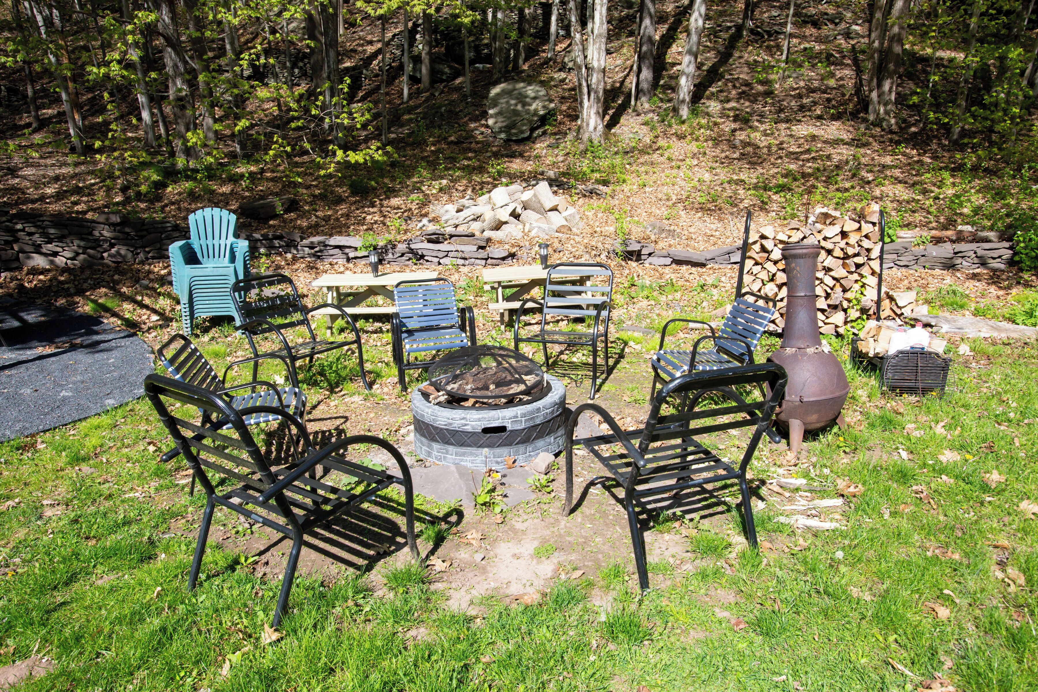 A circular seating area with a fire pit, surrounded by metal chairs and tables, set in a wooded area with a stone wall and stacked firewood in the background.