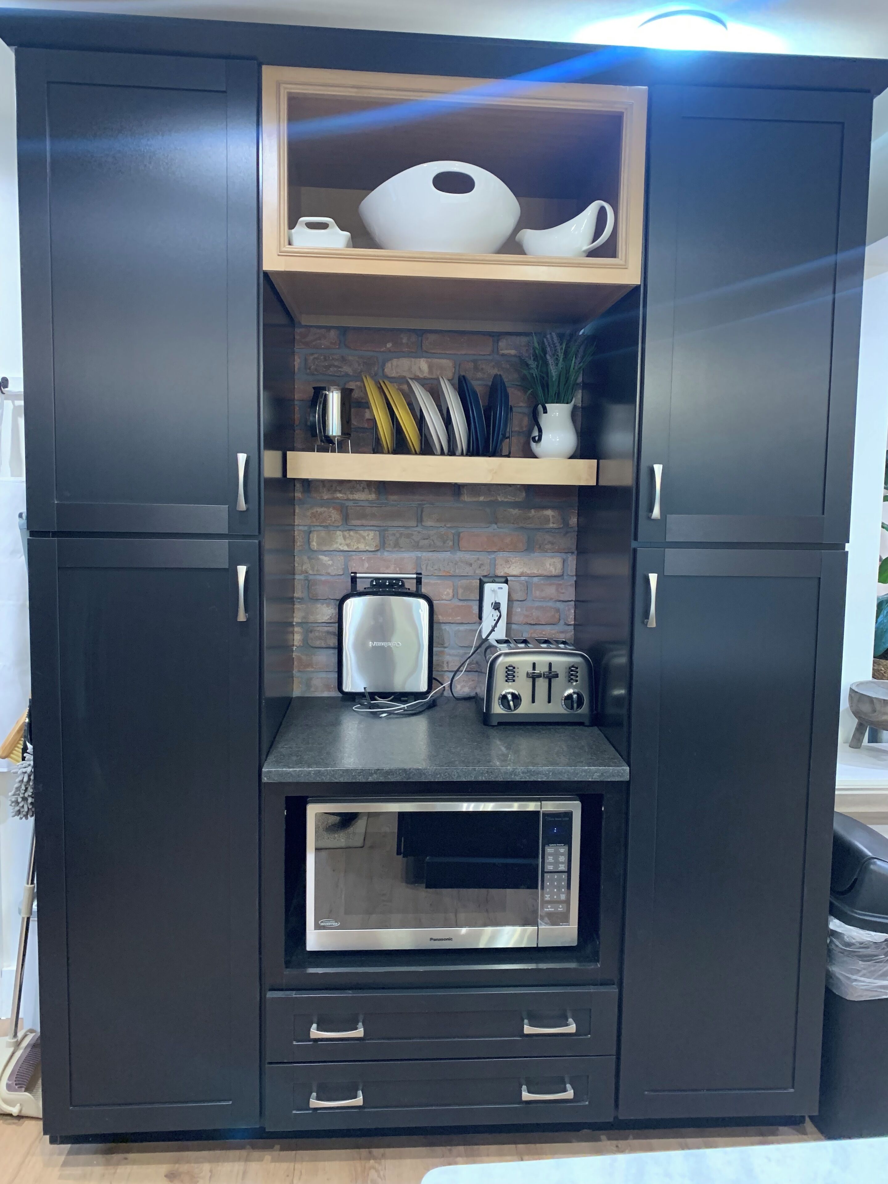 A kitchen with dark cabinetry, a microwave, a toaster, and a shelf with decorative plates and a potted plant.