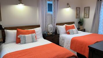 Une chambre d’hôtel avec deux lits, chacun agrémenté de draps blancs et orange, une tête de lit en bois, une table de chevet avec une lampe et une fenêtre avec des rideaux blancs.
