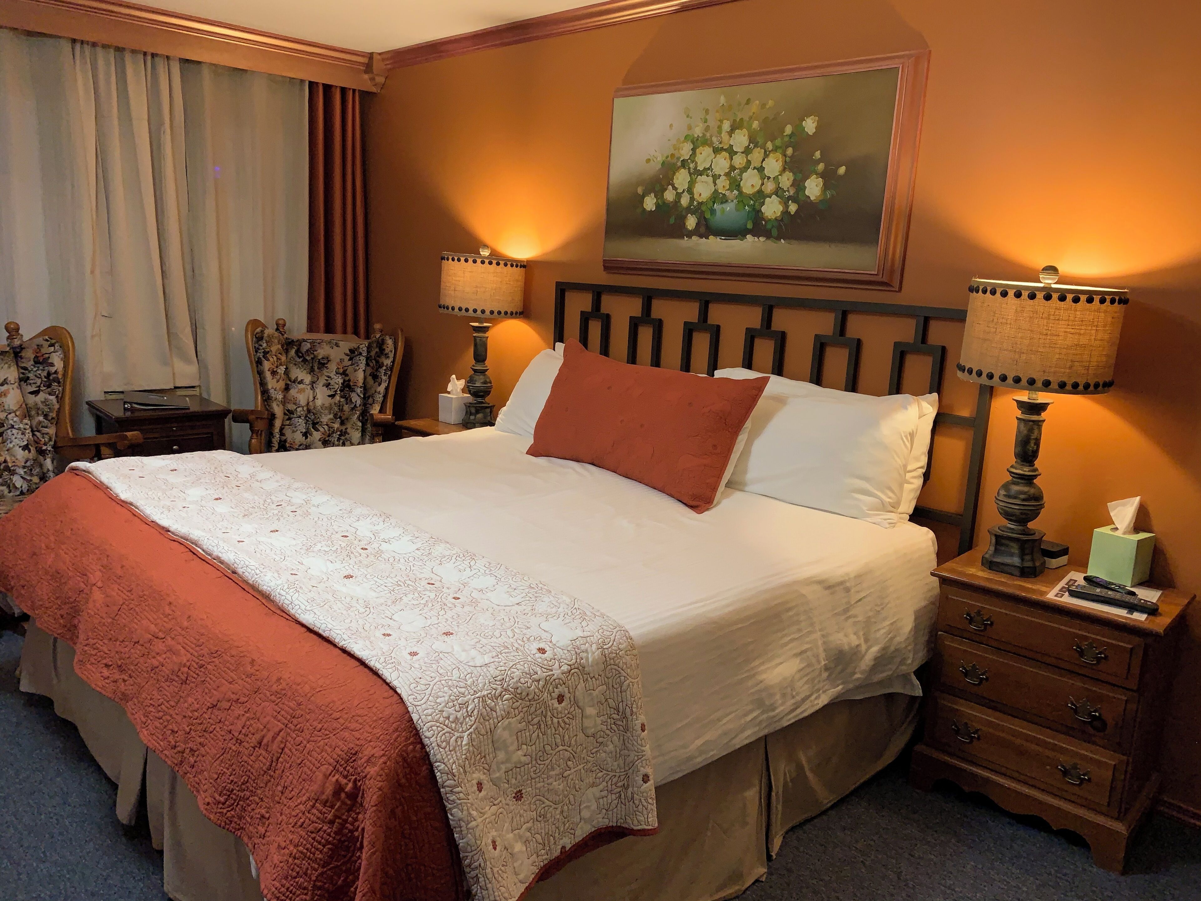 A neatly made bed with a patterned bedspread, two bedside lamps, a nightstand with a tissue box, and a painting above the bed.