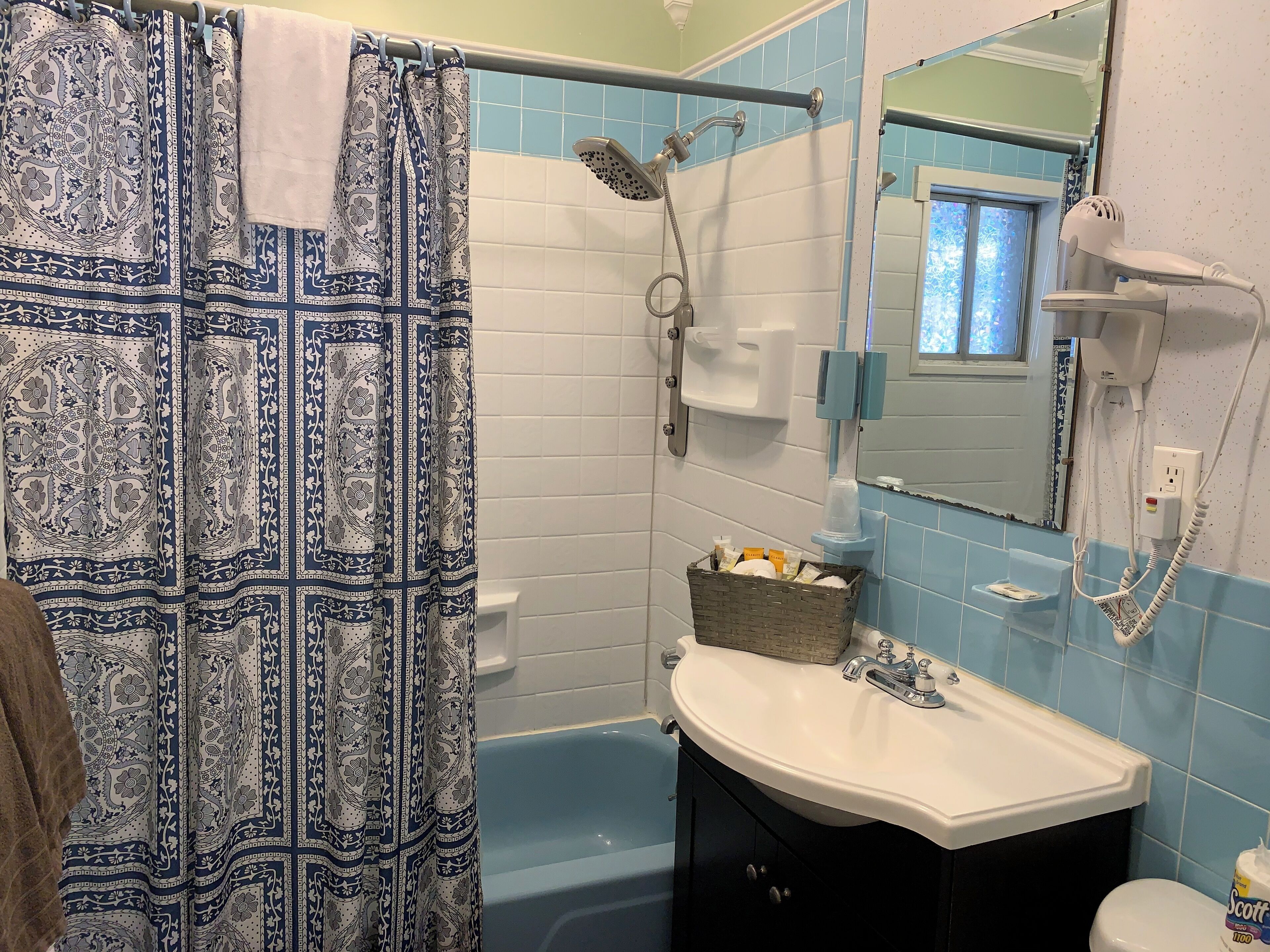 Une salle de bain avec un rideau de douche à motifs, un lavabo blanc, un miroir, une douchette à main et un panier contenant des articles de toilette.