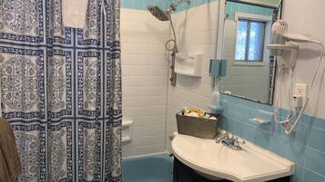 Une salle de bain avec un rideau de douche à motifs, un lavabo blanc, un miroir, une douchette à main et un panier contenant des articles de toilette.