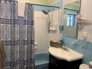 A bathroom with a patterned shower curtain, a white sink, a mirror, a handheld showerhead, and a basket of toiletries. - Starlite Motel (Big Indian)