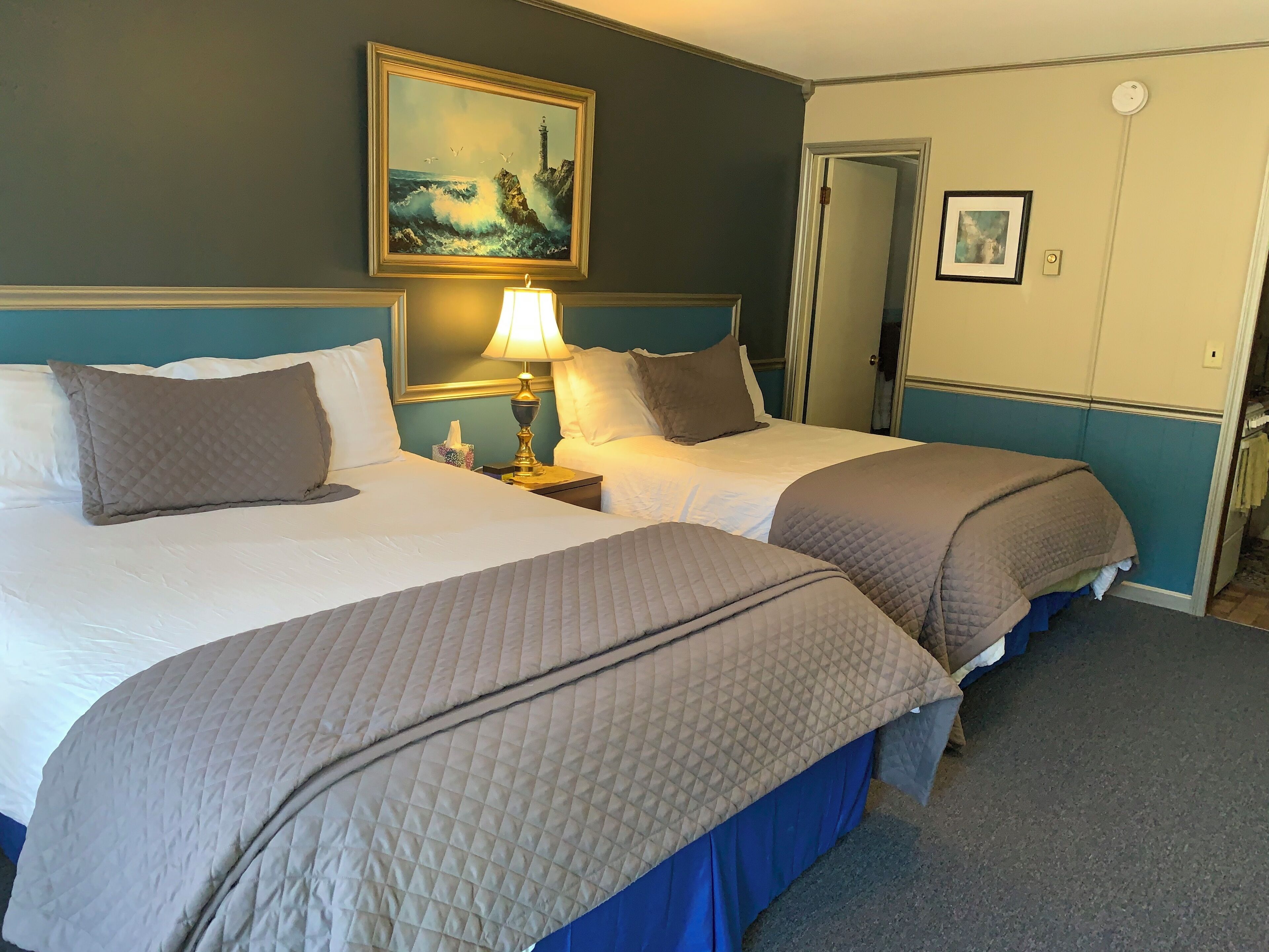 Habitación de hotel con dos camas, un cuadro de un faro, una lámpara de noche y una puerta que da a otra habitación.