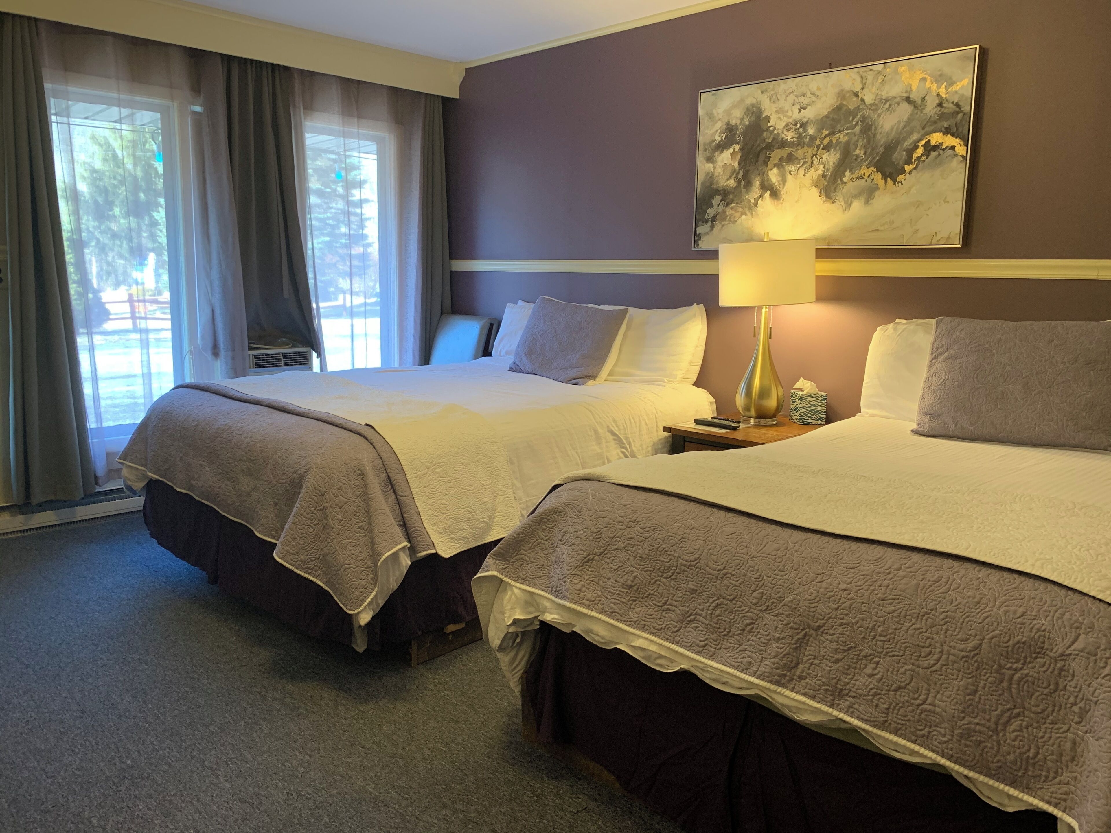 A hotel room with two beds, a large window with curtains, a painting on the wall, and a bedside lamp.